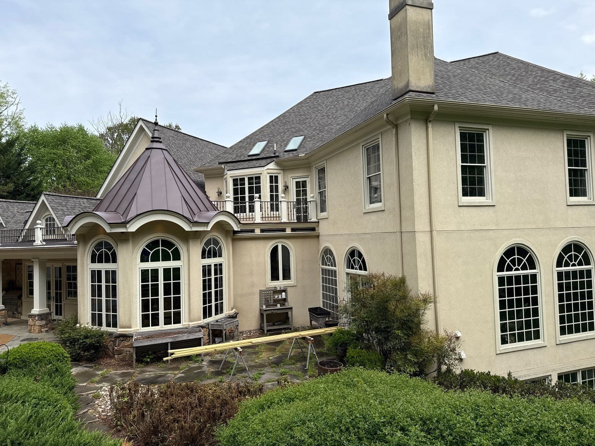 Large cream-colored house with distinctive octagonal sunroom featuring arched windows and metal roof, with construction materials in yard and mature landscaping